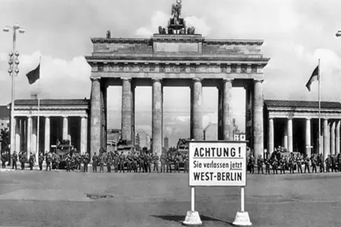 Brandenburger Tor, Soldaten und Panzer stehen Spalier. Ein Schild warnt: "ACHTUNG! Sie verlassen jetzt WEST-BERLIN". Ein Symbol des kalten Krieges.