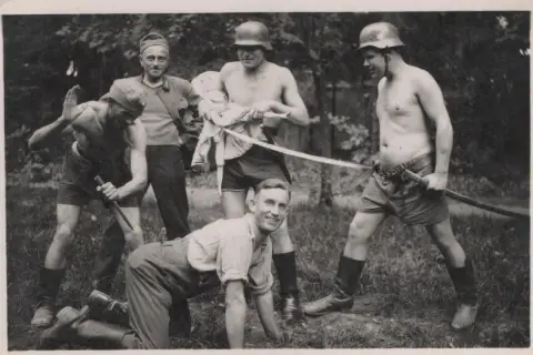 Fünf Männer, einige mit Helmen, posieren mit Waffen. Ein Mann hält eine Puppe. Die Szene erinnert an die Verbrechen der Wehrmacht.