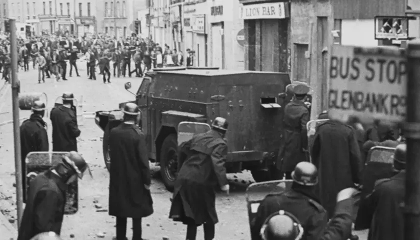 Unruhen in Nordirland, ein gepanzertes Fahrzeug und Polizisten mit Schilden. Die Szene erinnert an die Zeit vor dem Karfreitagsabkommen.