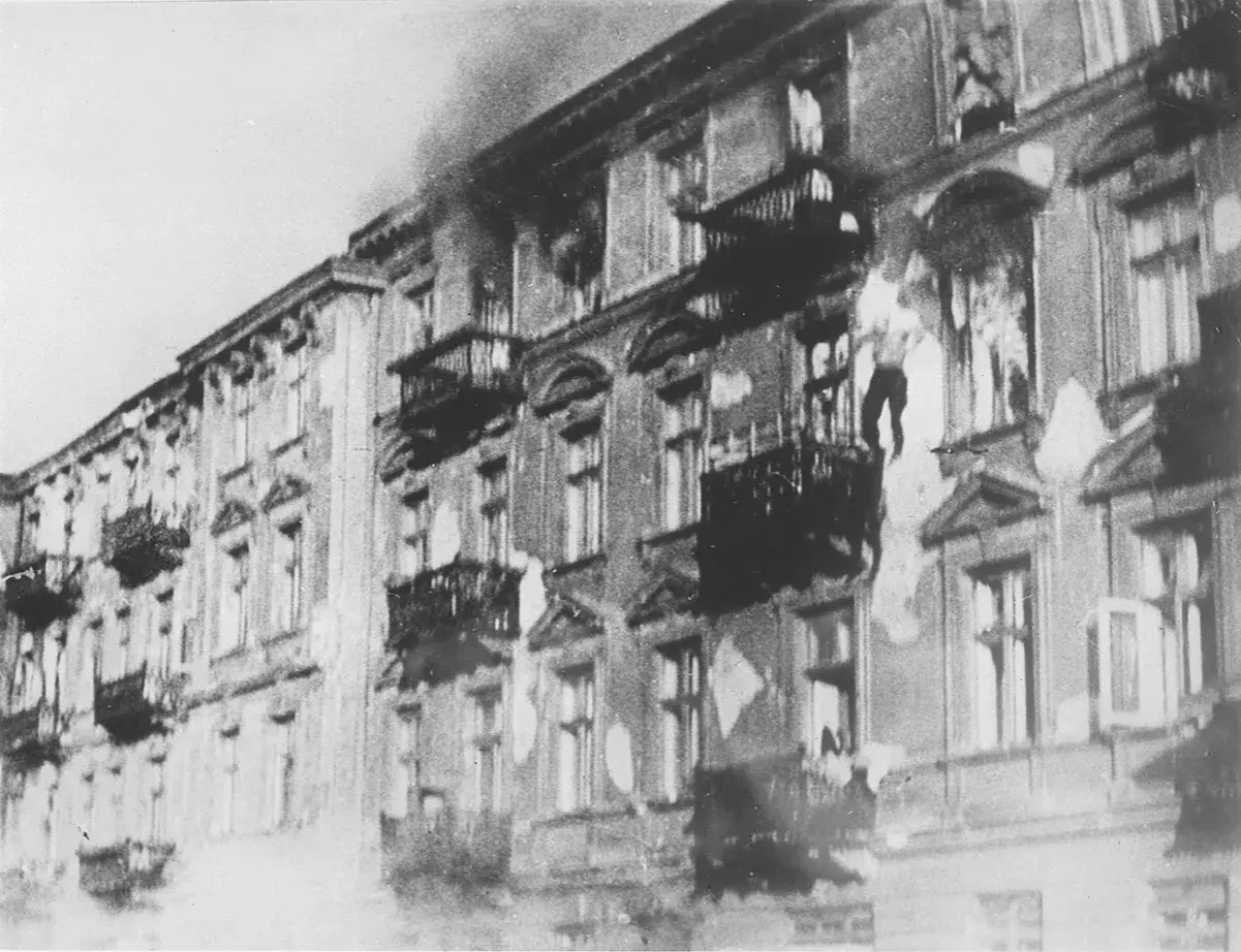 Brennende Gebäude im Warschauer Ghetto. Rauch steigt auf, Fenster sind zerbrochen, Balkone sind dunkel.