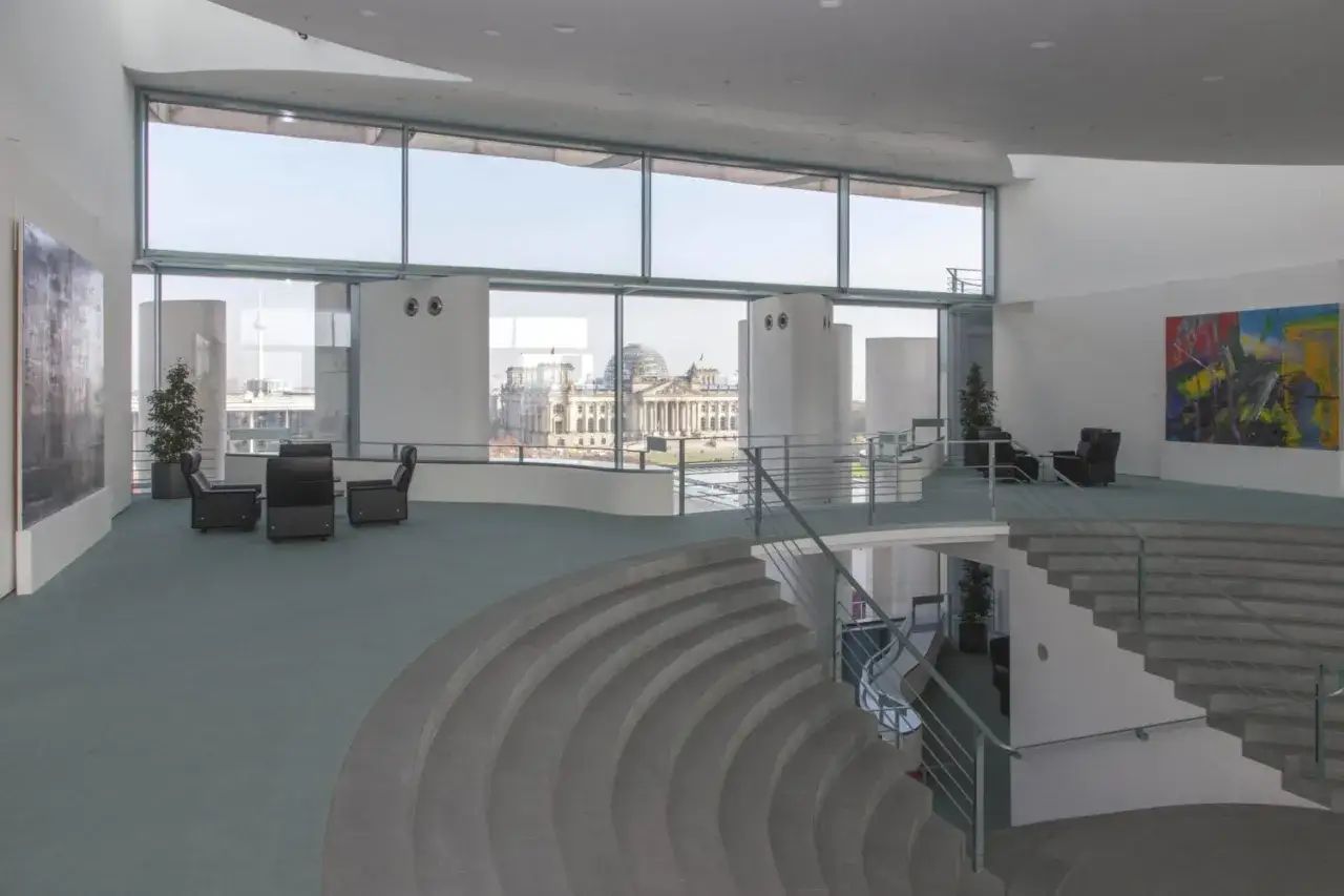 Modernes Foyer im Bundeskanzleramt innen mit Blick auf das Reichstagsgebäude. Sitzgruppen und eine geschwungene Treppe prägen den Raum.