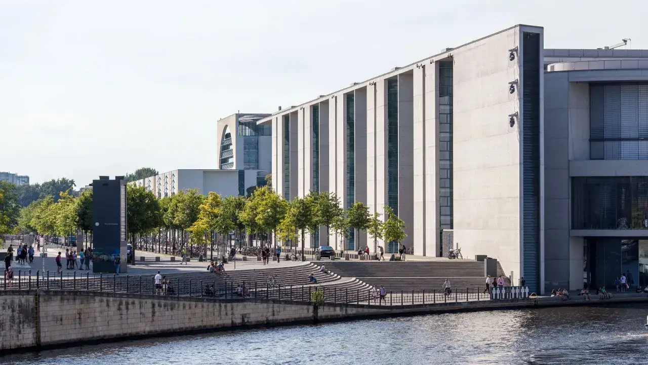 Moderne Architektur am Spreeufer in Berlin, die an die Grenzen der Macht des Bundeskanzlers erinnert. Menschen flanieren, sitzen und genießen die Sonne.
