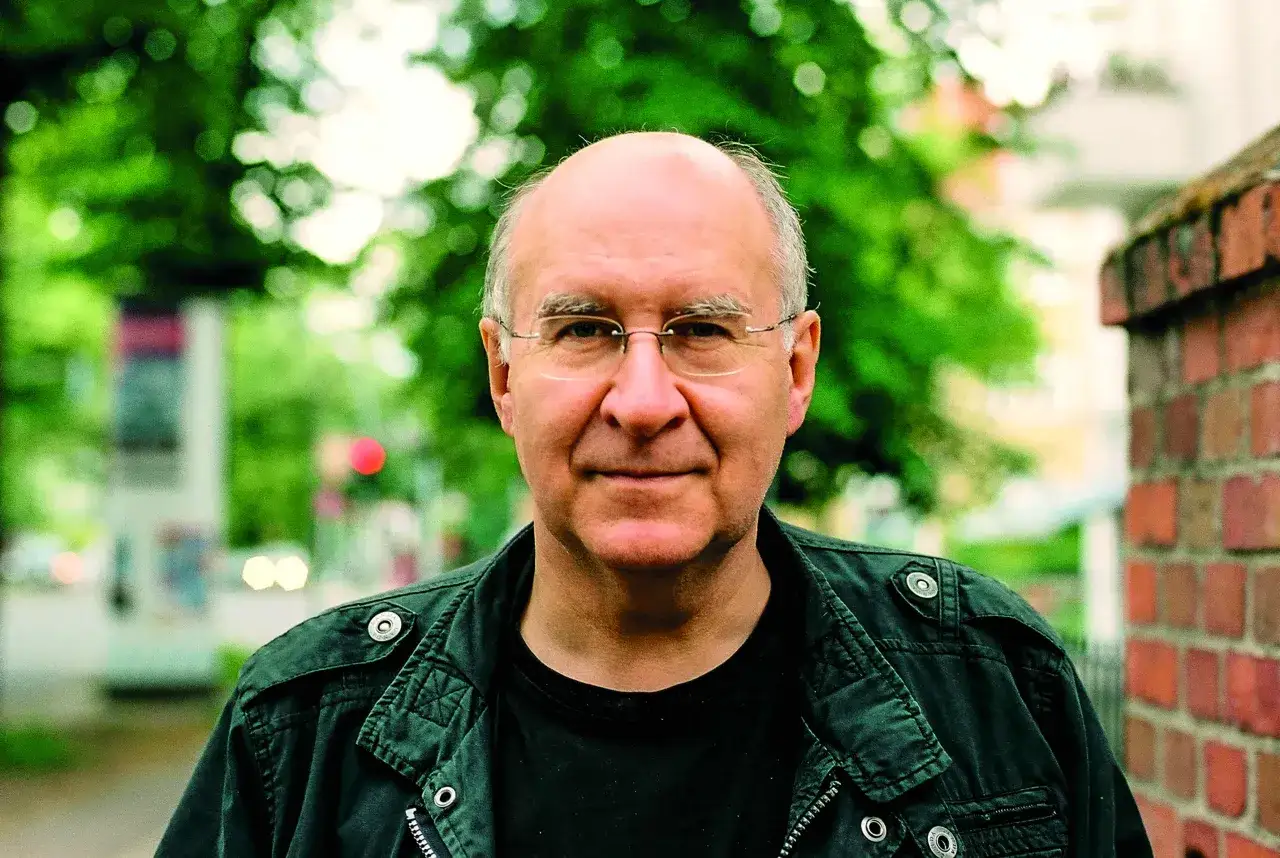 Portr&auml;t von Peter Wensierski, einem Mann mit Brille und Glatze, der eine dunkle Jacke tr&auml;gt. Im Hintergrund sind B&auml;ume und eine Ziegelmauer zu sehen.