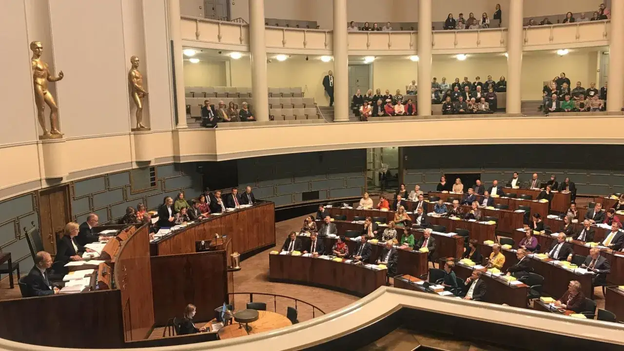 Sitzung im finnischen Parlament. Abgeordnete sitzen in hölzernen Pulten, während Statuen die Wände schmücken.
