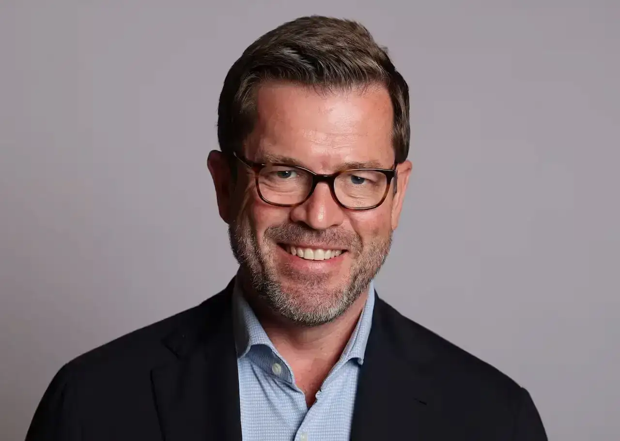 Porträt von Karl-Theodor zu Guttenberg mit Brille und Anzug, lächelnd vor grauem Hintergrund.