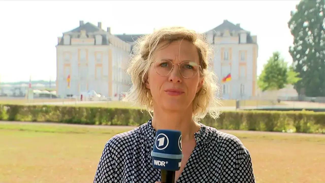 Eine Reporterin hält ein Mikrofon vor einem Schloss, auf dem die Flaggen von Deutschland und Frankreich wehen.