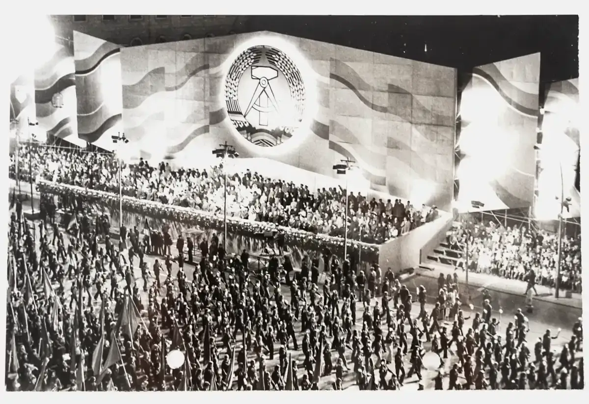 Große Parade am 7. Oktober, dem Tag der Republik DDR. Tausende marschieren mit Fahnen vor einer riesigen Bühne mit dem Emblem der DDR.