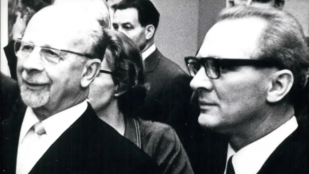 Zwei M&auml;nner mit Brillen, einer mit Bart, im Gespr&auml;ch. Der Mann links k&ouml;nnte ein DDR Chef sein.