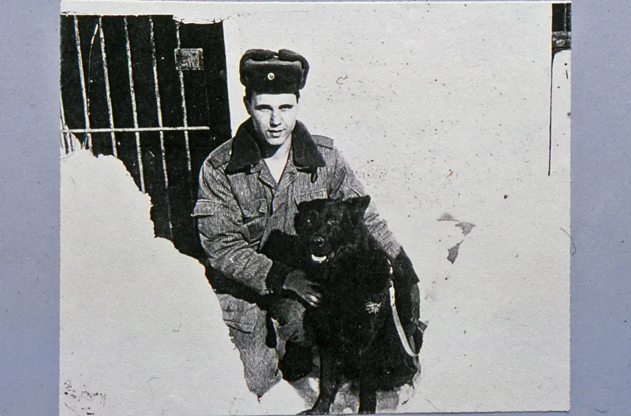 Ein junger Mann, Dietmar Schultke, in Uniform und mit Mütze, sitzt im Schnee und hält einen schwarzen Hund.